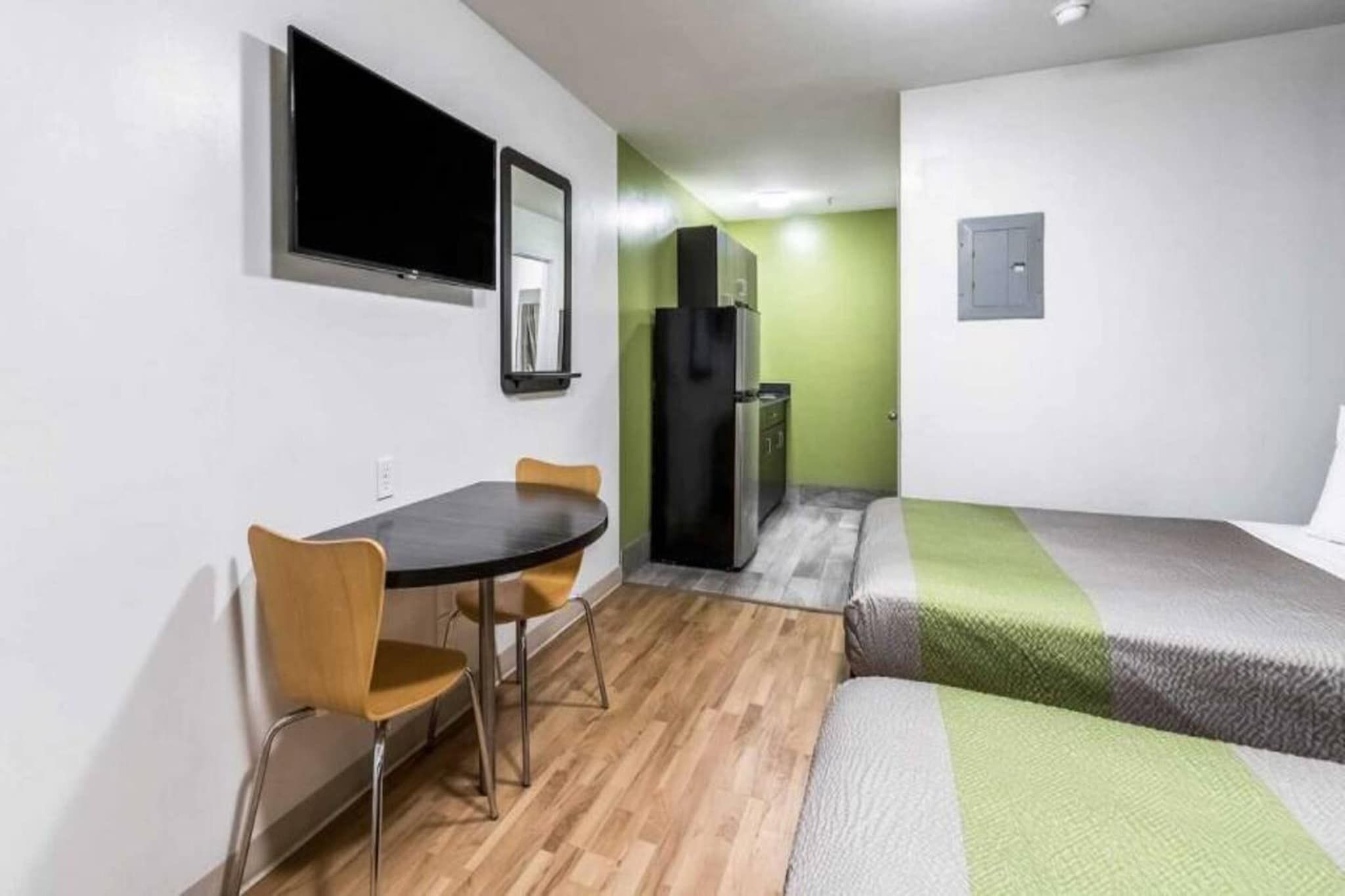 A compact room with a bed, a small table with chairs, a television mounted on the wall, and a refrigerator.