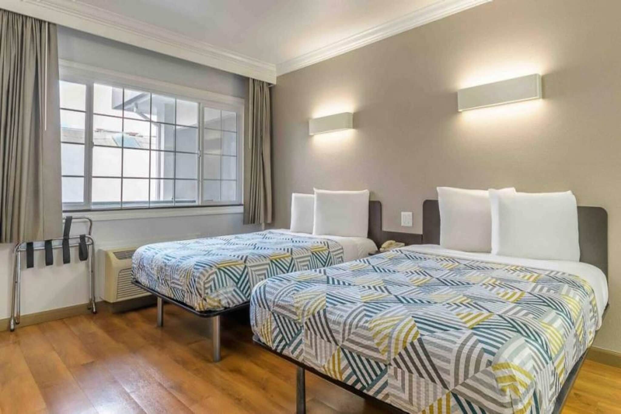 Quarto de hotel com duas camas, janela com cortinas, mesa de cabeceira com abajur e radiador.