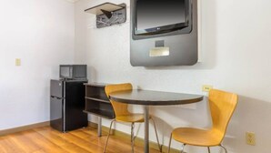 Habitación compacta con televisión de pantalla plana en la pared, un refrigerador pequeño y una mesa redonda con dos sillas.