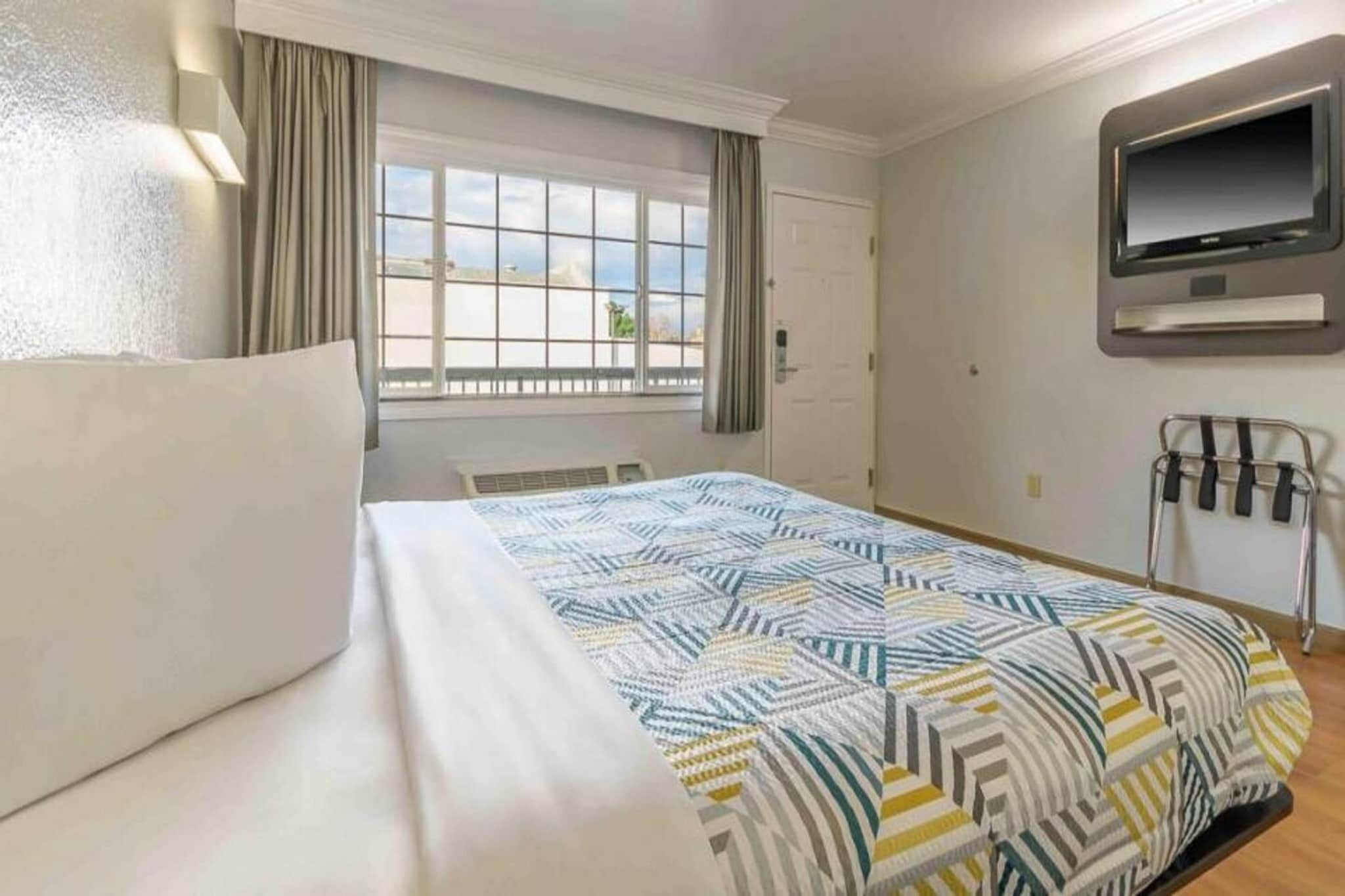 Habitación de hotel con una cama, un televisor montado en la pared y un ventanal con cortinas.