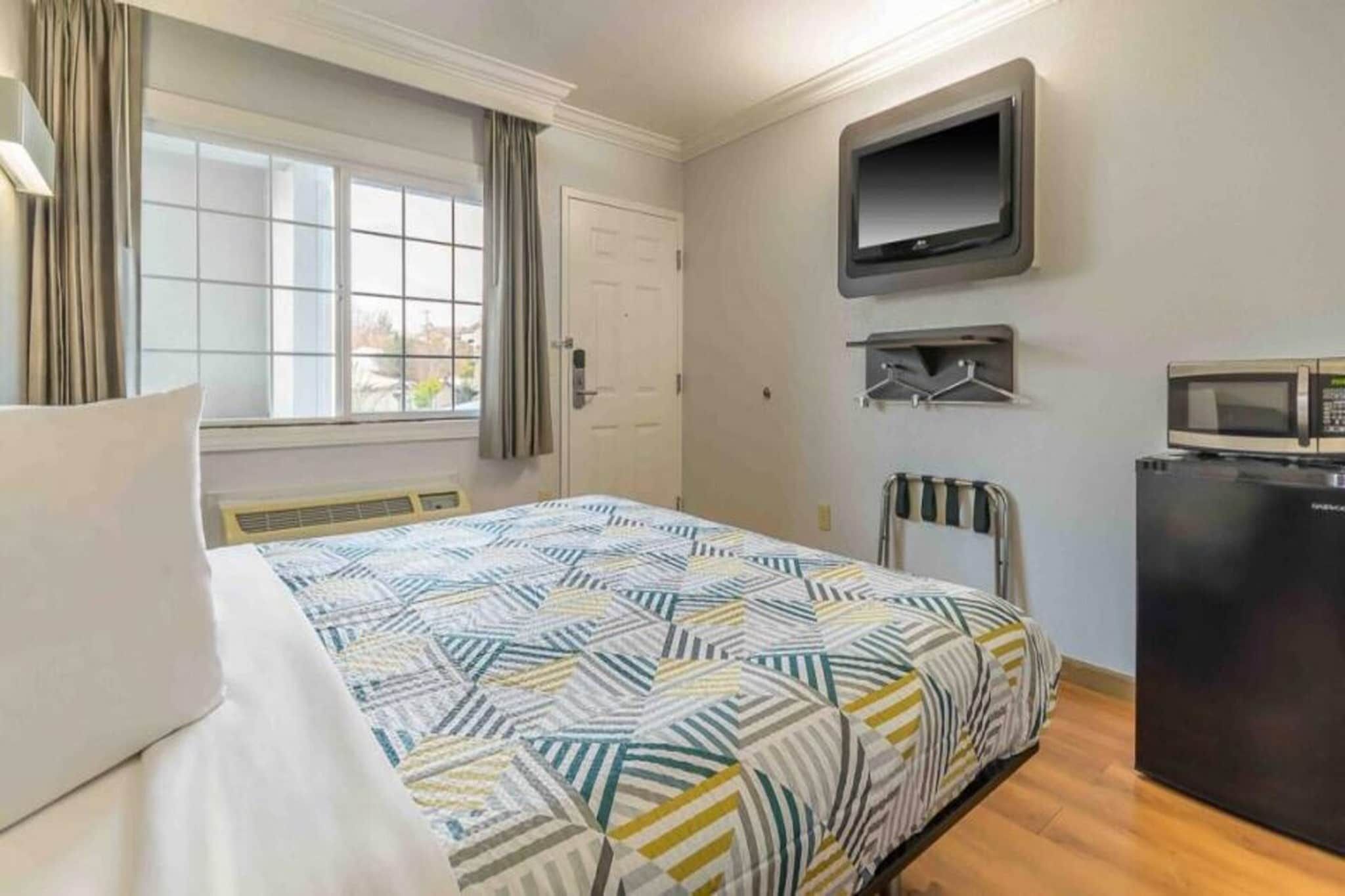 Habitación de hotel con cama, televisor montado en la pared, escritorio pequeño con microondas y ventana con cortinas.