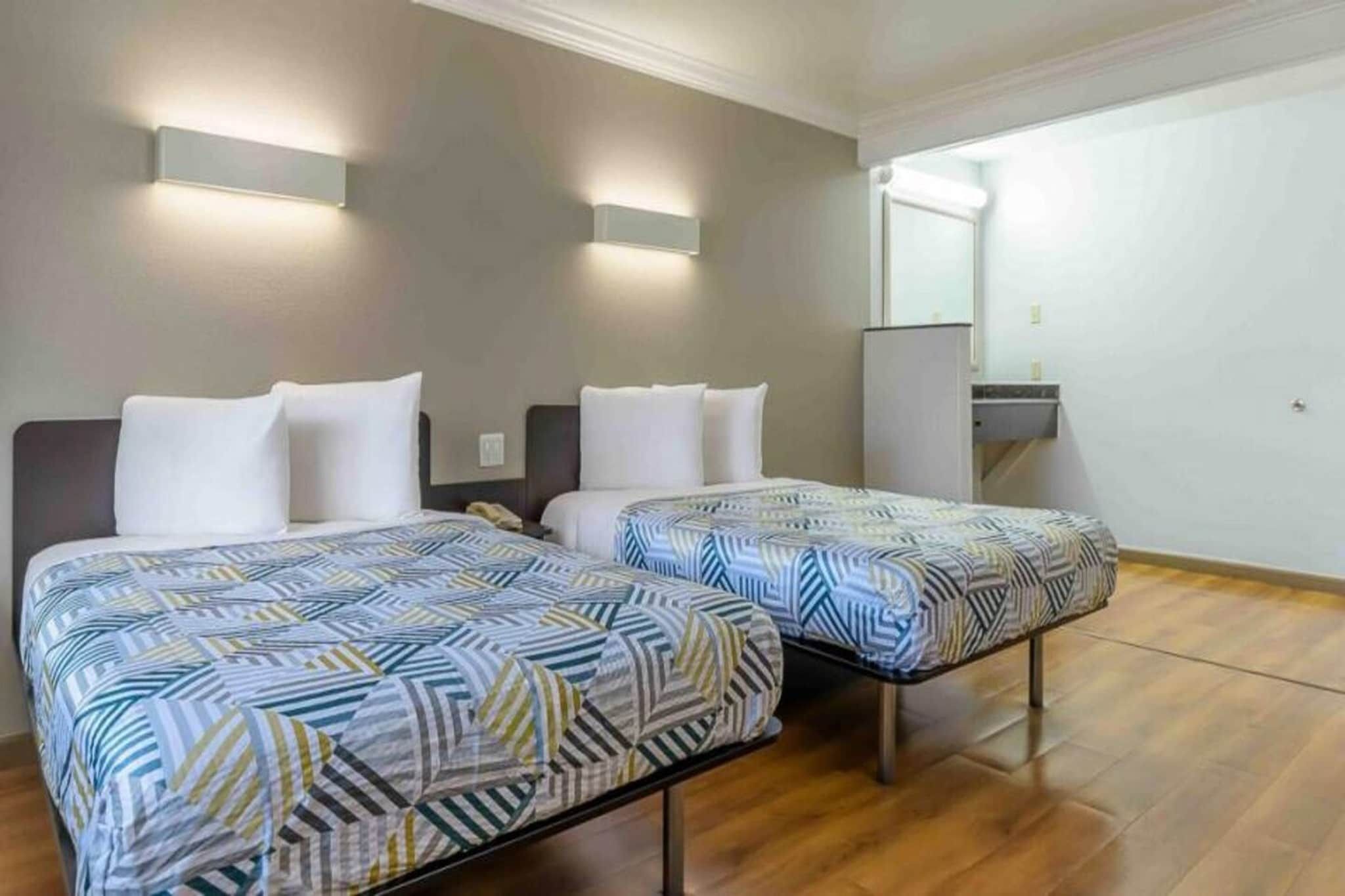 A hotel room with two beds, each with a patterned bedspread, a bedside table, a nightstand, and a wall-mounted light fixture.
