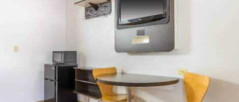 Una habitación pequeña con una mesa redonda y dos sillas, un televisor montado en la pared y un refrigerador.