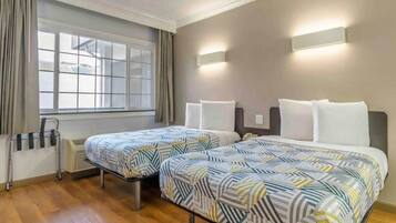 A hotel room with two beds, a window with curtains, a bedside table with a lamp, and a radiator.