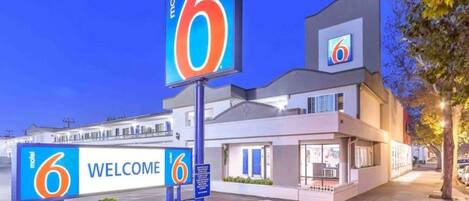 Un motel con un llamativo letrero con el número '6', un cartel de 'BIENVENIDOS' y una fachada de edificio moderna.