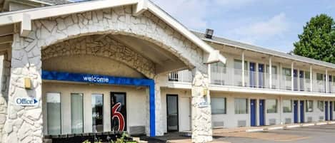 A motel with a stone arch entrance, a welcome sign, and a blue '6' sign indicating room number.