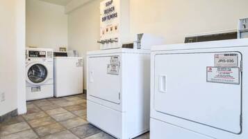 A laundry room with a front-loading washer and dryer, a wall-mounted soap and bleach dispenser, and a sign indicating the location of the restrooms.