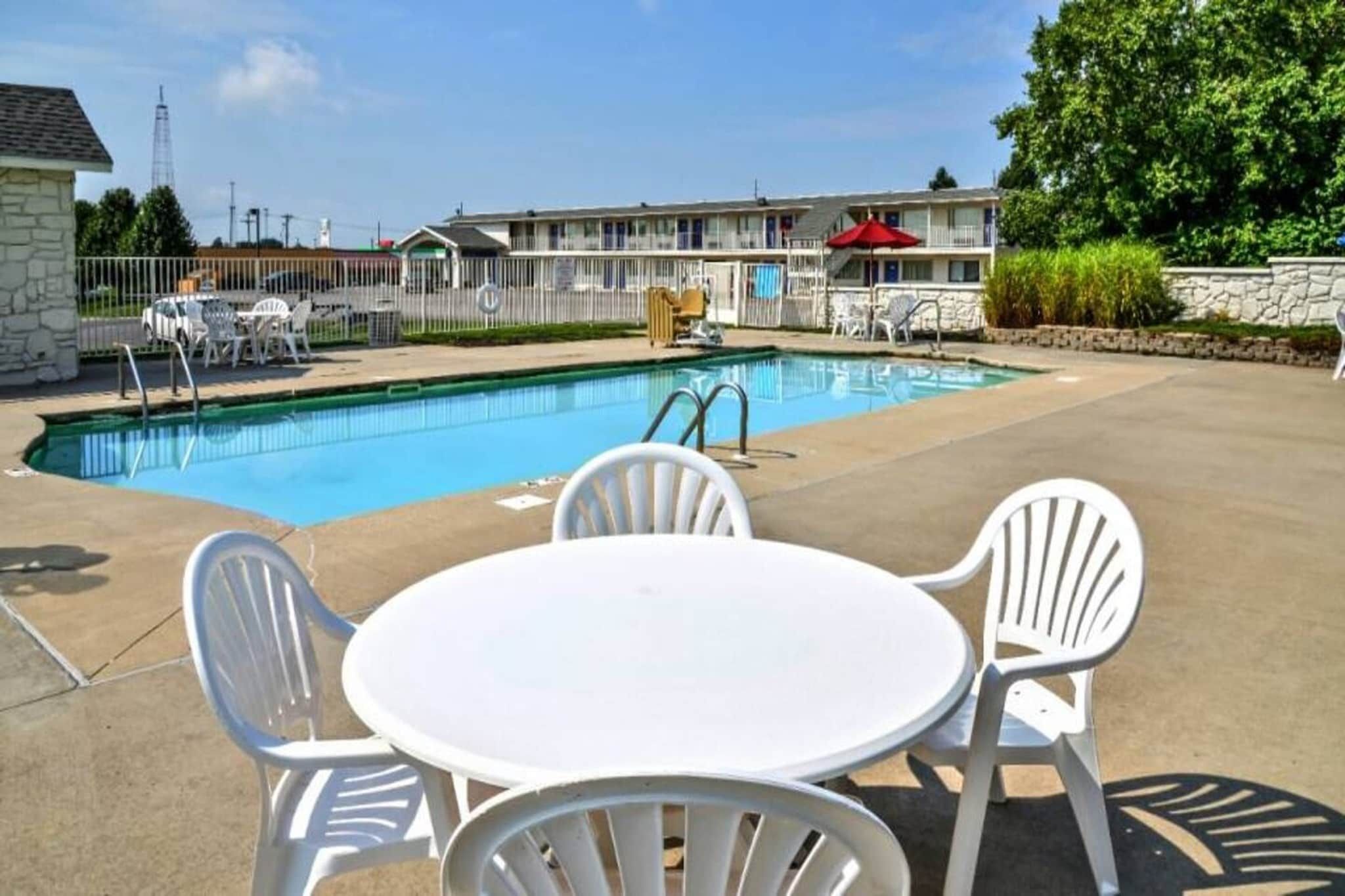 Una piscina con zona de patio, que incluye una mesa y sillas, y un edificio de motel al fondo.