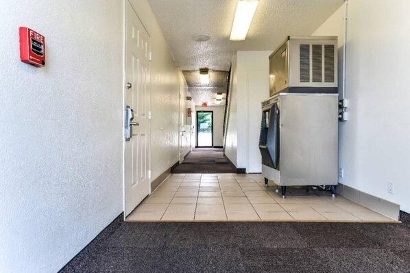 A hallway with a fire alarm on the left, a water fountain on the right, and a door at the end.