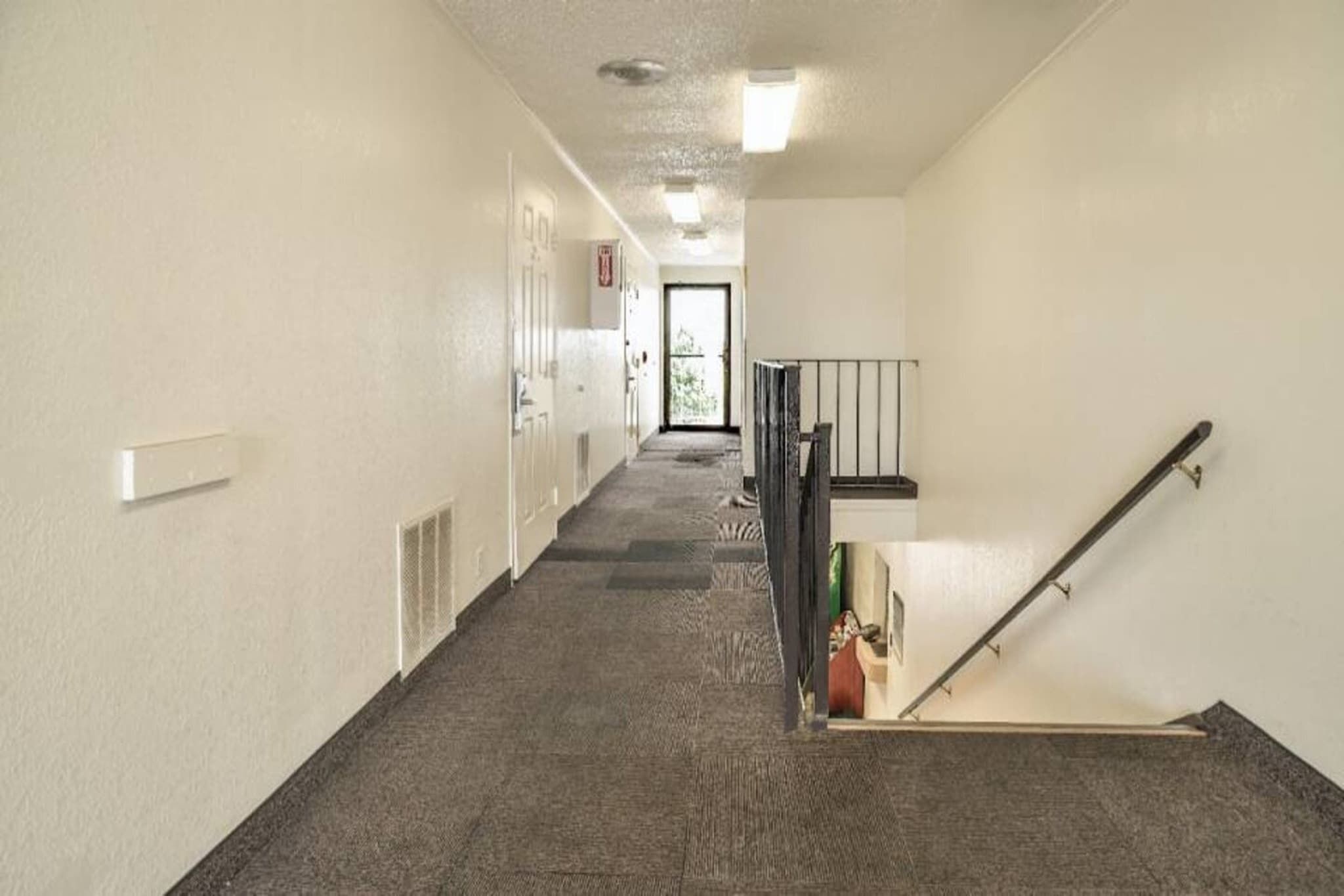 A hallway with carpeted flooring, a door at the end, and a staircase with a handrail.