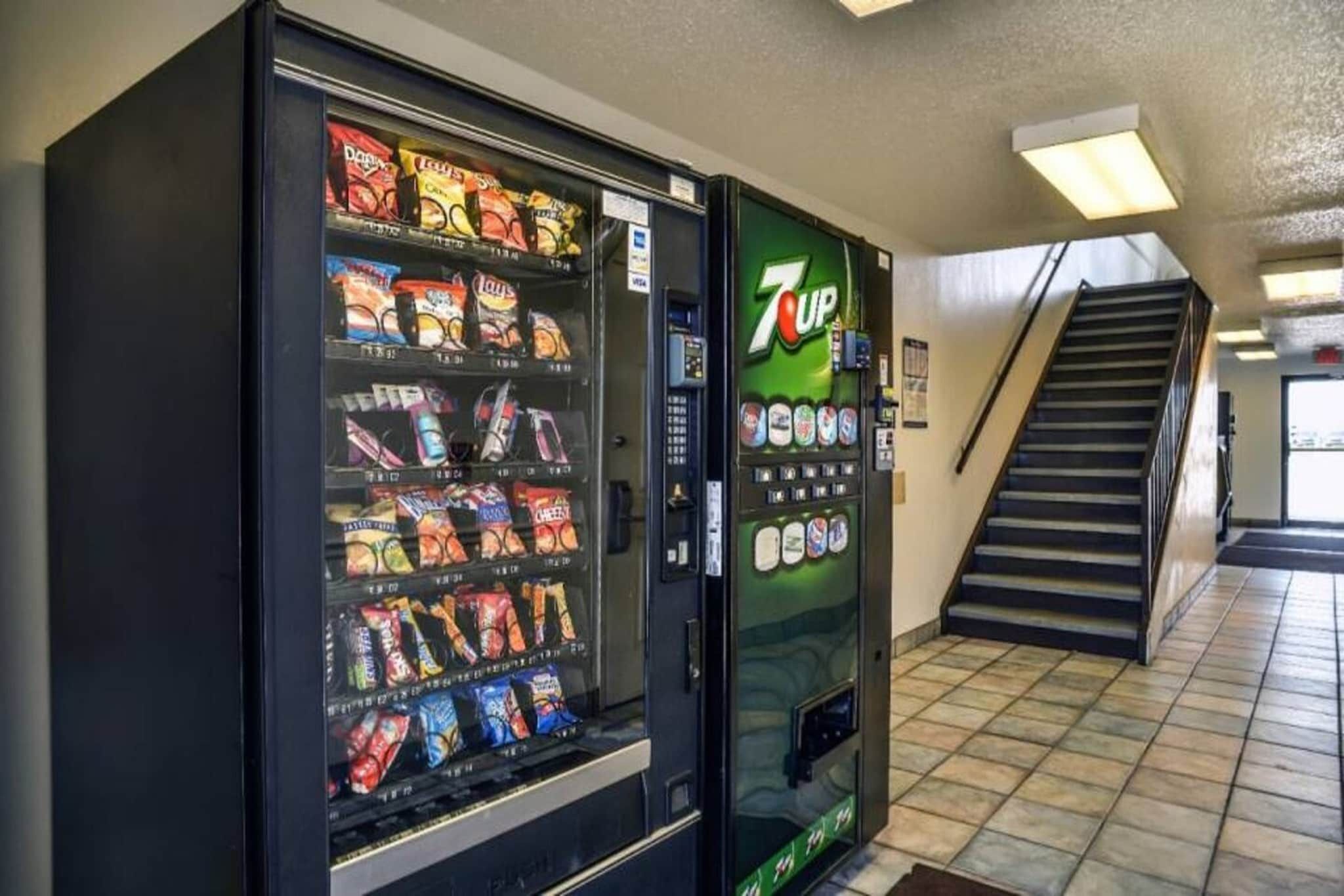 Máquina de vendas com lanches e dispensador de refrigerante 7UP em um corredor com escada.