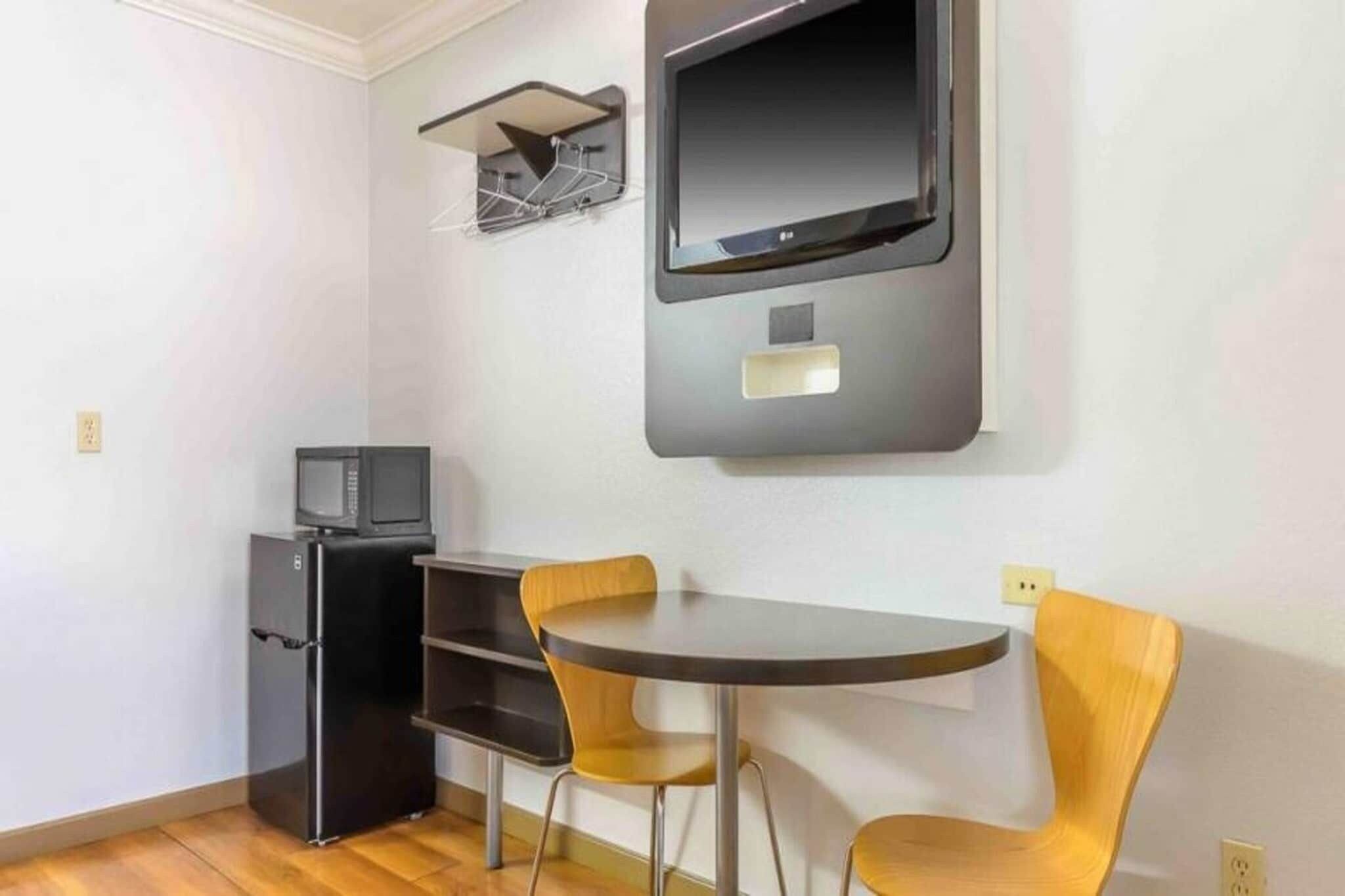 A compact room with a small refrigerator, a microwave, a flat-screen TV mounted on the wall, and a round table with two chairs.