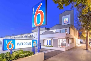 Un motel con un llamativo letrero con el número '6', un cartel de 'BIENVENIDOS' y una fachada de edificio moderna.