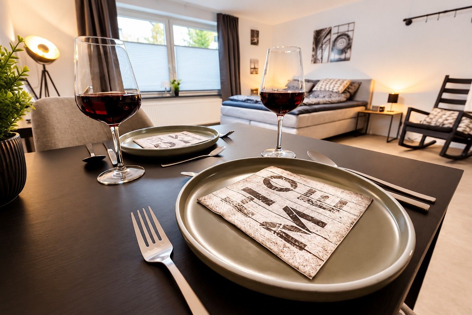 Ein Esstisch ist gedeckt mit Tellern, Gläsern und Besteck in einem modernen Wohnzimmer mit einer Couch und einer Lampe.