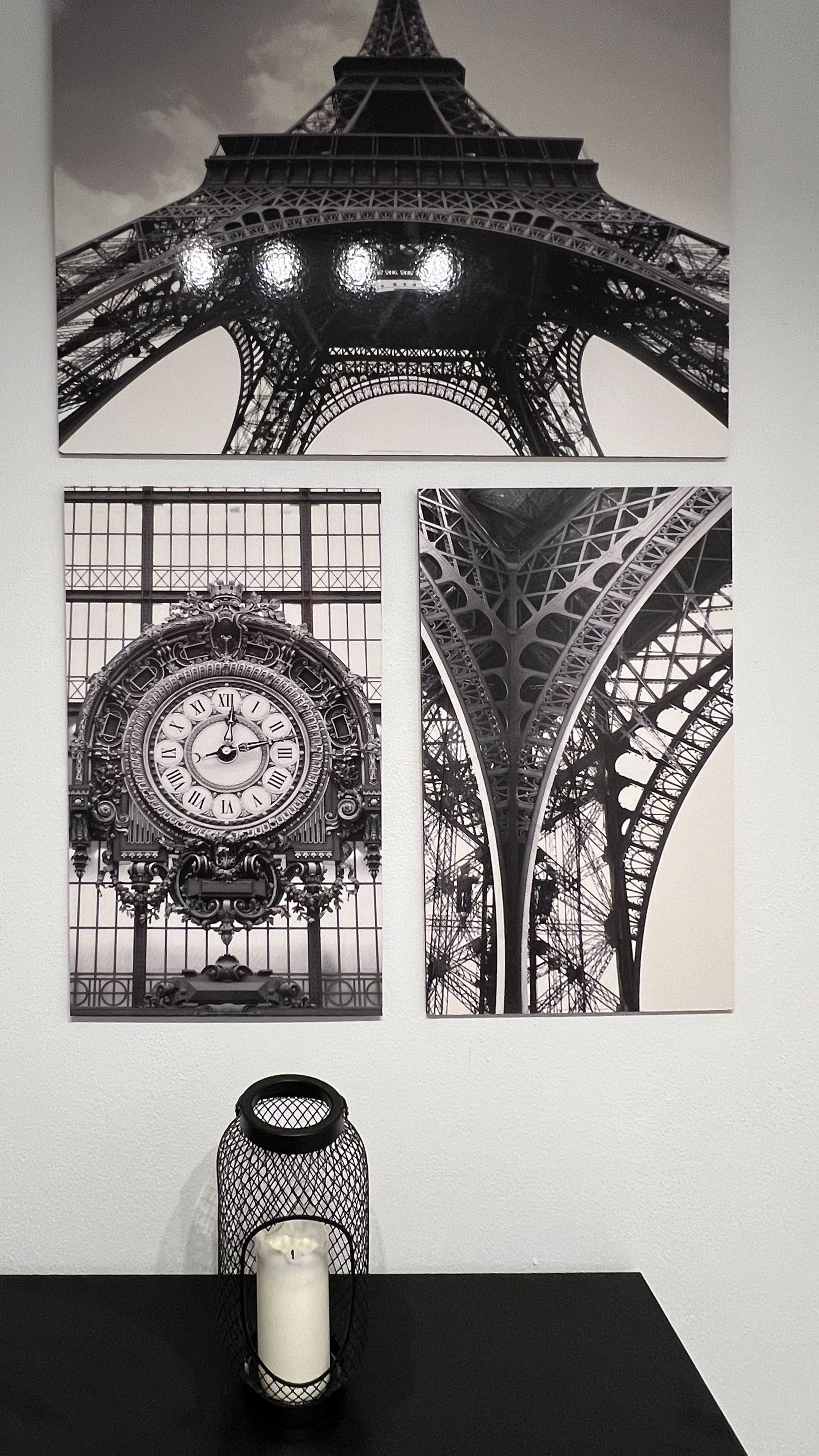 A wall with three black and white photographs: a clock, the Eiffel Tower, and a bridge. Below, a candle in a wire holder is visible.