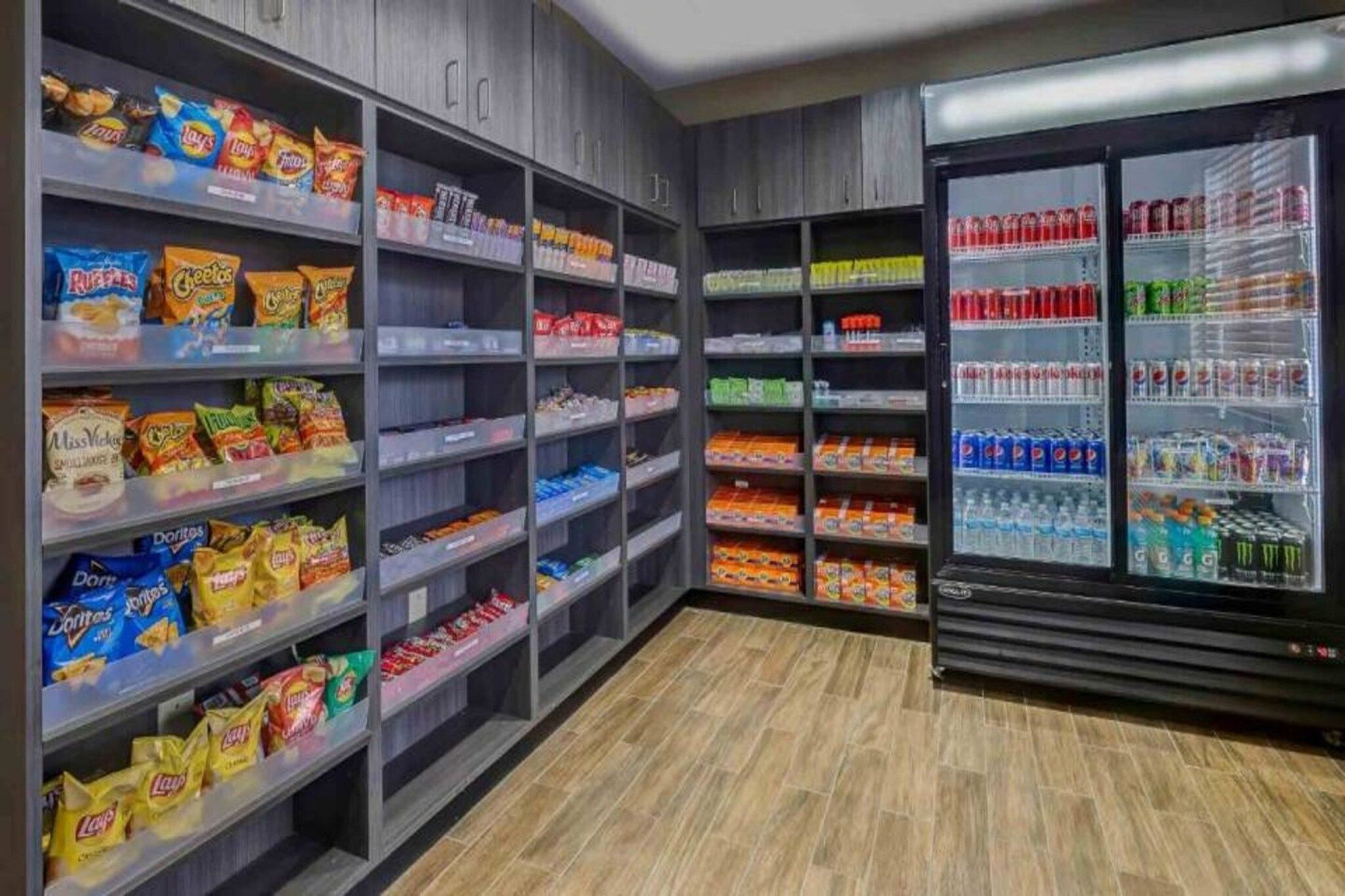 Uma área de lanches e bebidas com prateleiras repletas de salgadinhos, biscoitos e um refrigerador com refrigerantes e água.