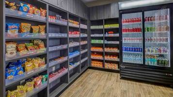 Uma área de lanches e bebidas com prateleiras repletas de salgadinhos, biscoitos e um refrigerador com refrigerantes e água.