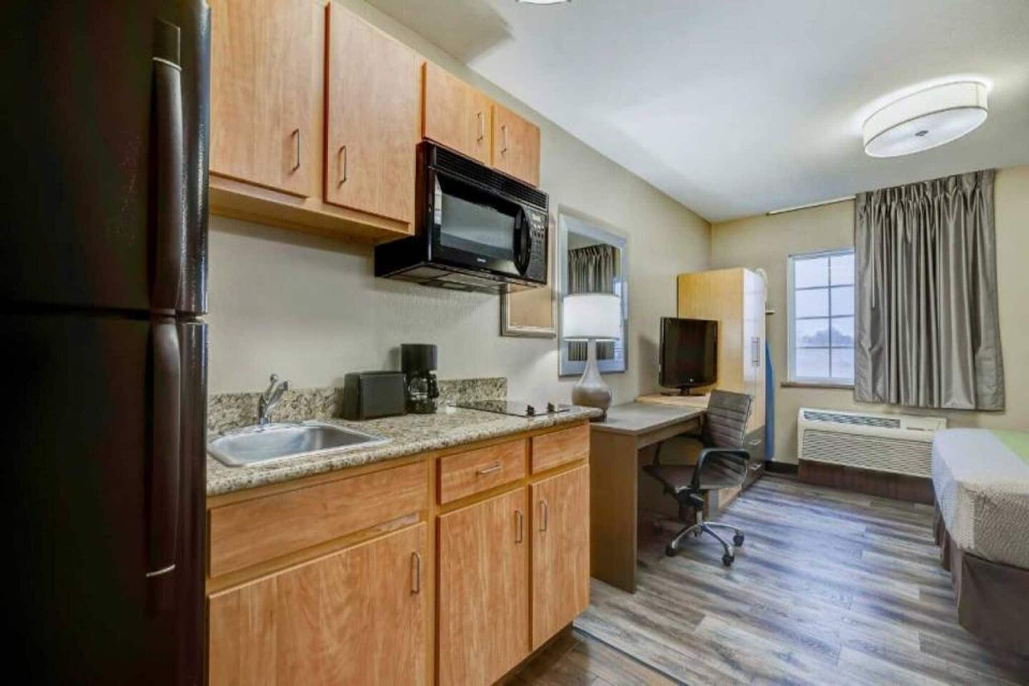 A compact hotel room with a kitchenette, a single bed, a desk with a chair, and a window with curtains.