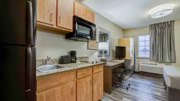 A compact hotel room with a kitchenette, a single bed, a desk with a chair, and a window with curtains.