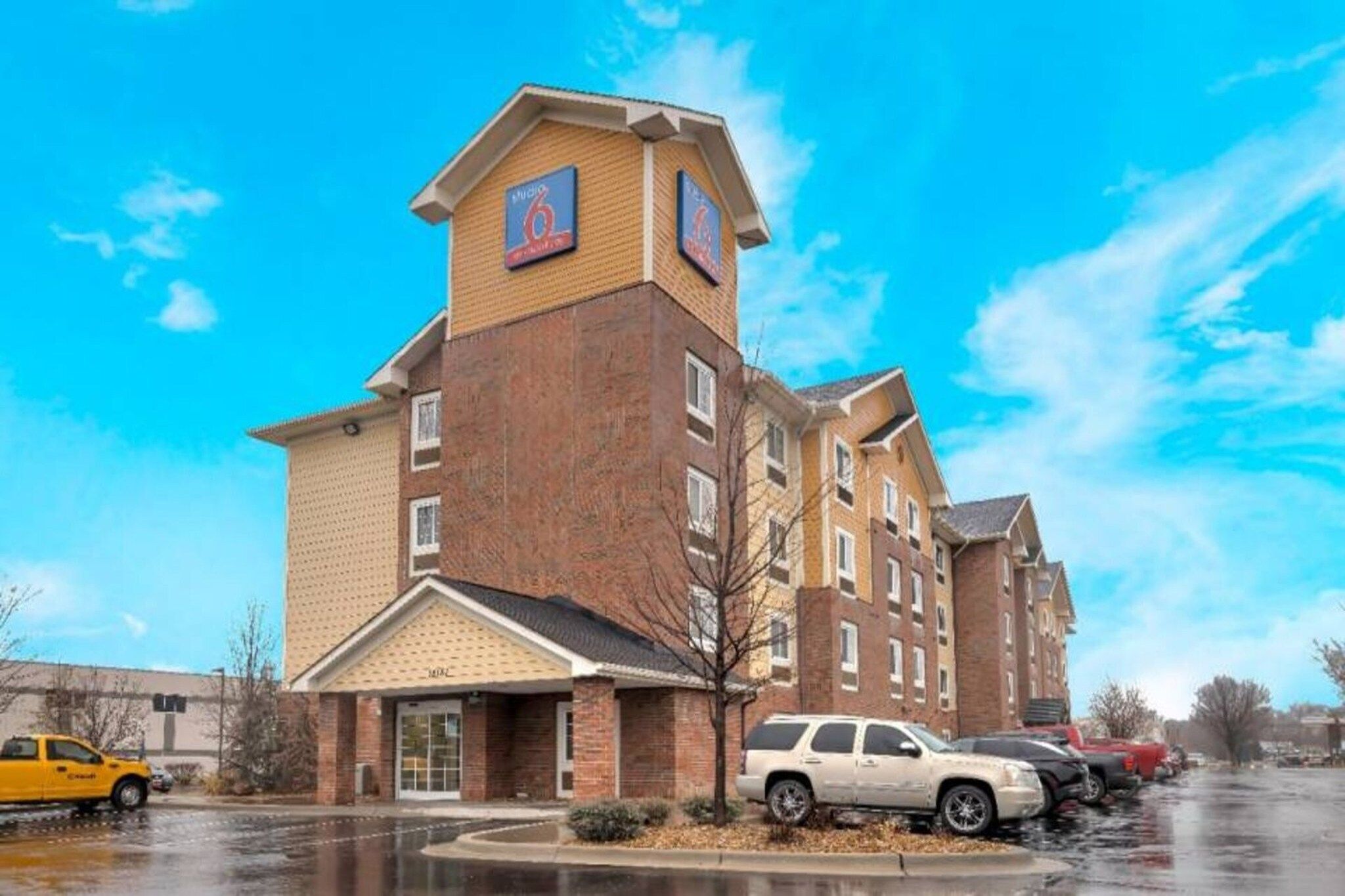 Un hôtel à plusieurs étages, doté d’une façade en briques, d’une entrée centrale et d’un parking où sont stationnés des véhicules.