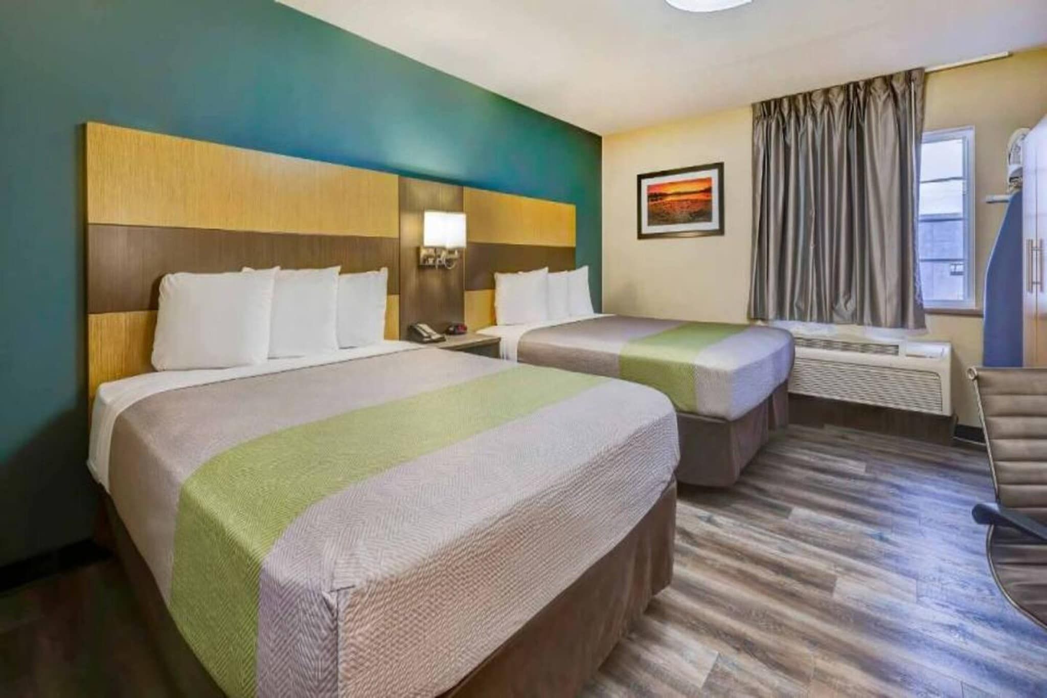 A hotel room with two beds, a nightstand with a lamp, a window with curtains, and a painting on the wall.