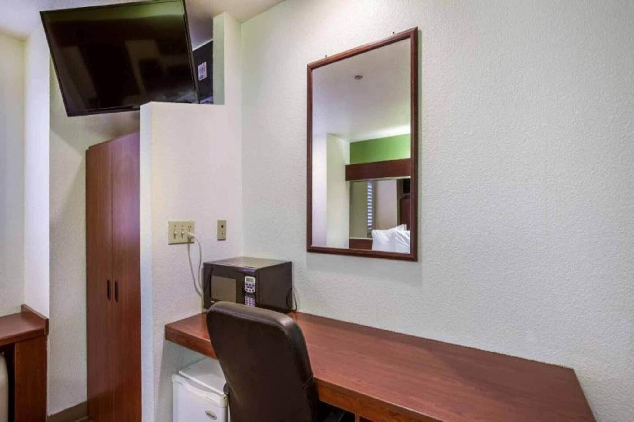 A hotel room with a wooden desk, a chair, a television mounted on the wall, a microwave, and a small refrigerator.