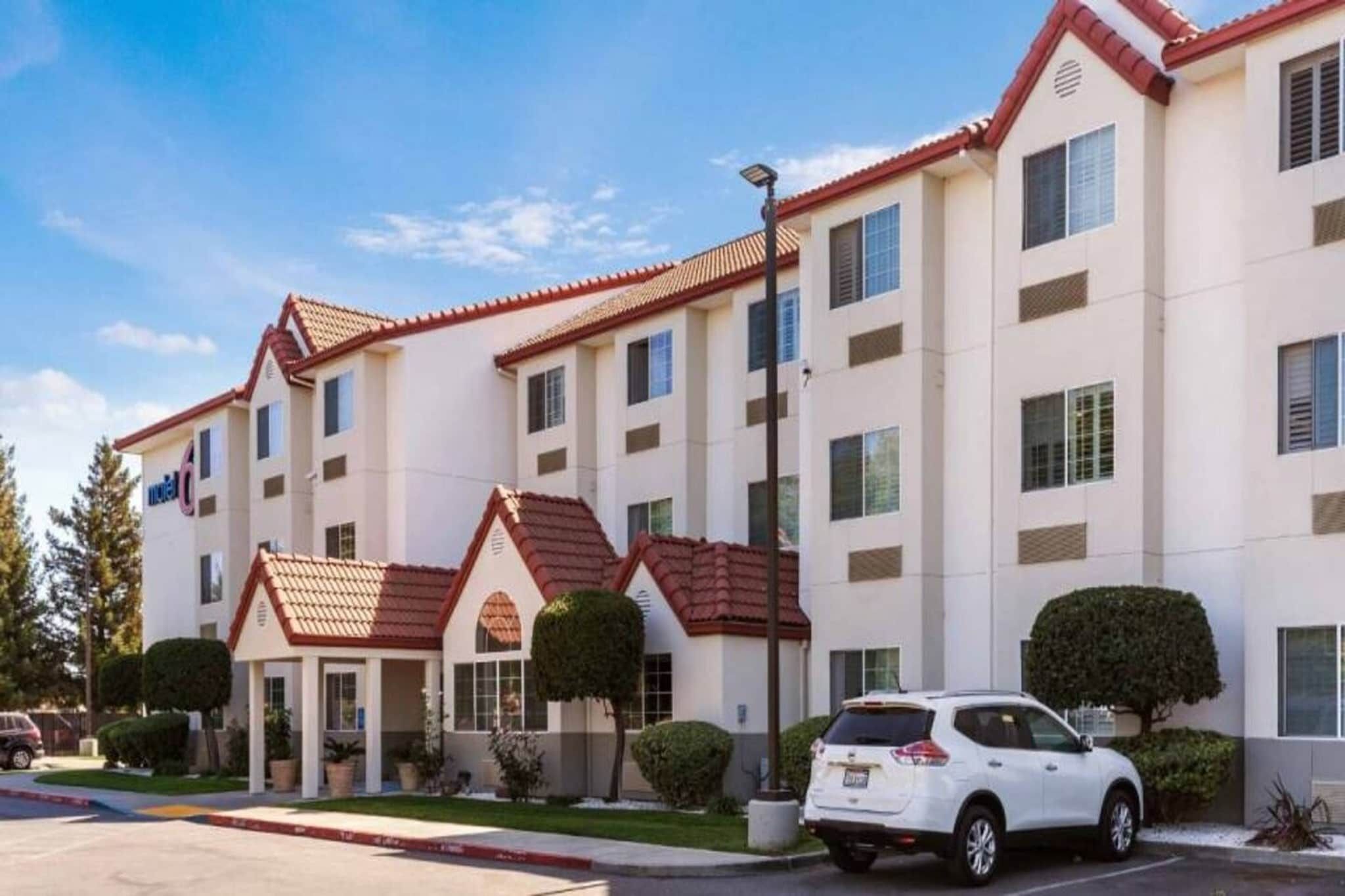 Un hôtel à plusieurs étages, avec un toit recouvert de tuiles rouges, un parking et une voiture blanche garée devant.