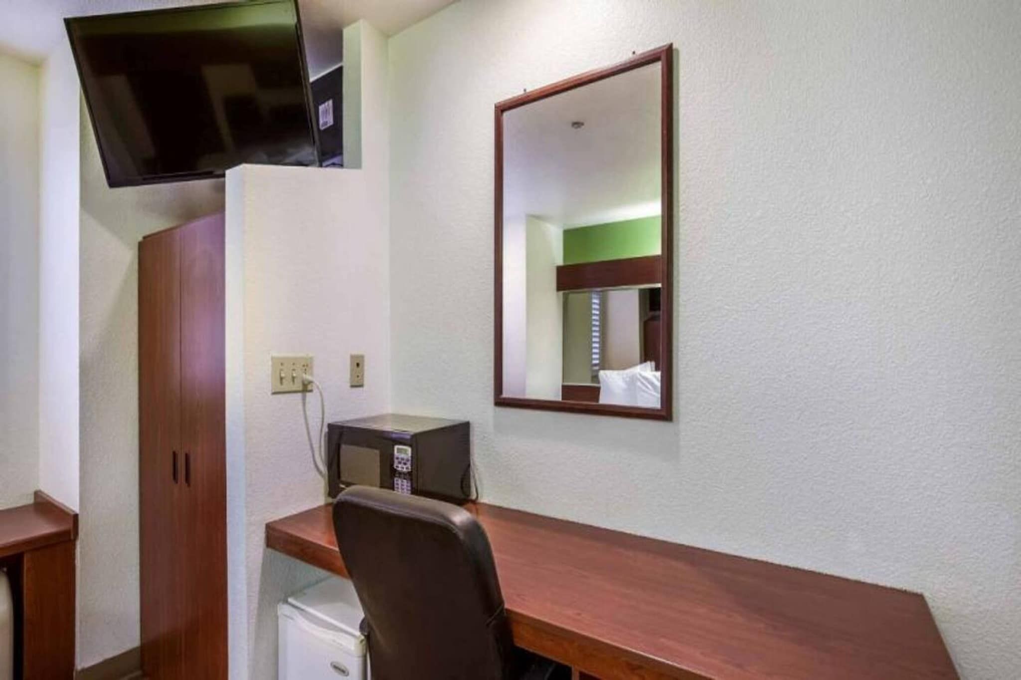 Ein Hotelzimmer mit einem Holztisch, einem Sessel, einem an der Wand befestigten Fernseher, einer Mikrowelle und einem kleinen Kühlschrank.