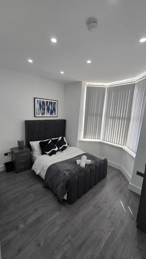 Un dormitorio con cama, mesitas de noche, una ventana con persianas y una lámpara de techo.