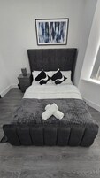 Una cama con cabecera gris, ropa de cama blanca y dos almohadas blanco y negro.