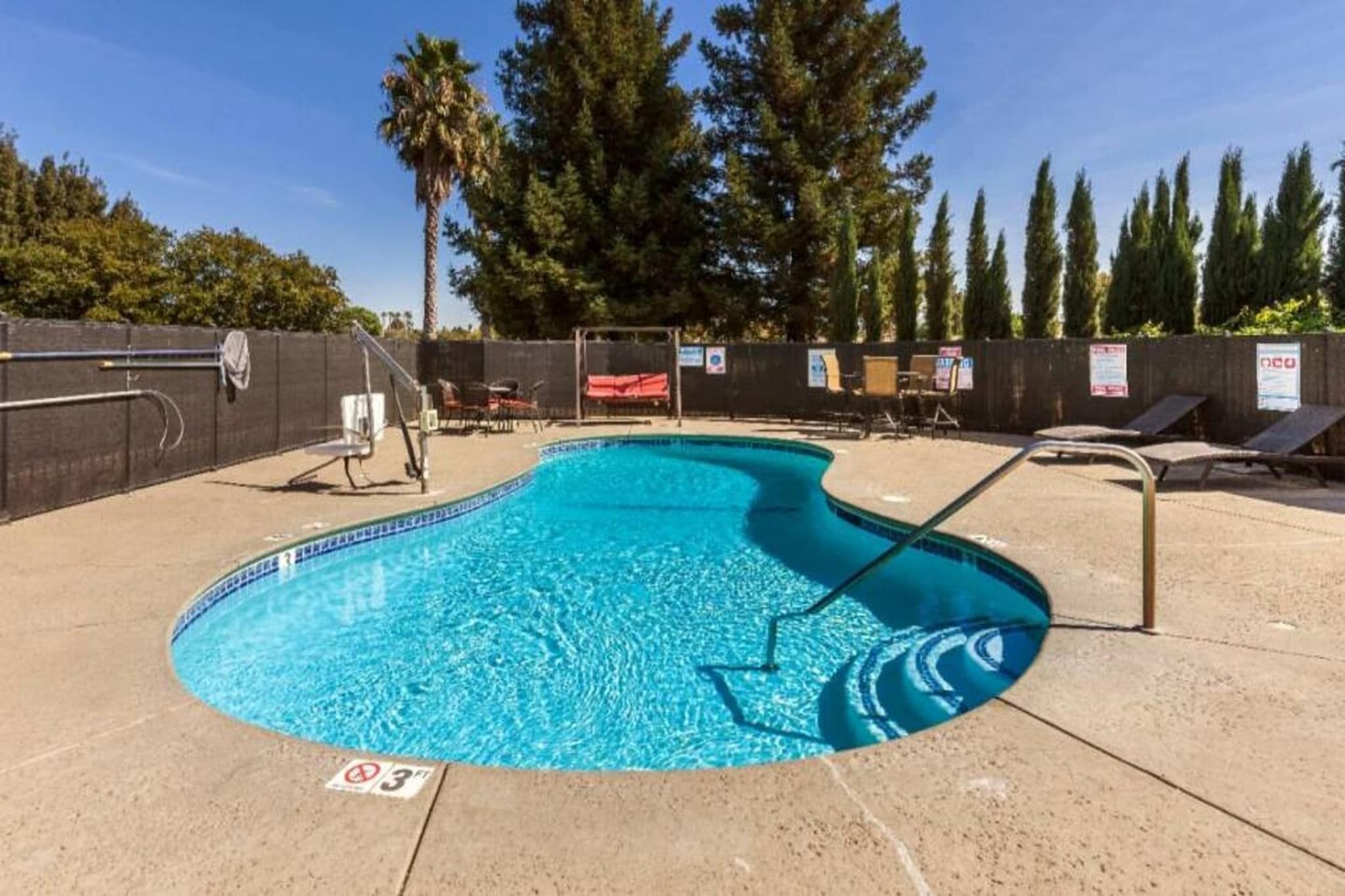 A swimming pool with a diving board, surrounded by lounge chairs and a fence.