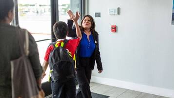 Une personne portant une chemise bleue et une veste noire traverse un seuil, accompagnée d’un enfant vêtu d’une chemise rouge et d’un short noir.