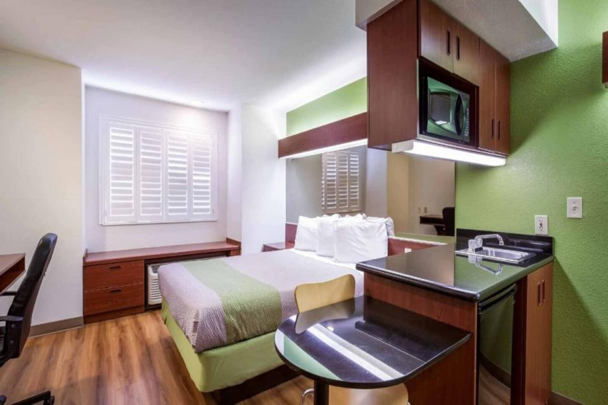 A compact hotel room with a bed, a desk, a chair, a small kitchenette, and a window with blinds.