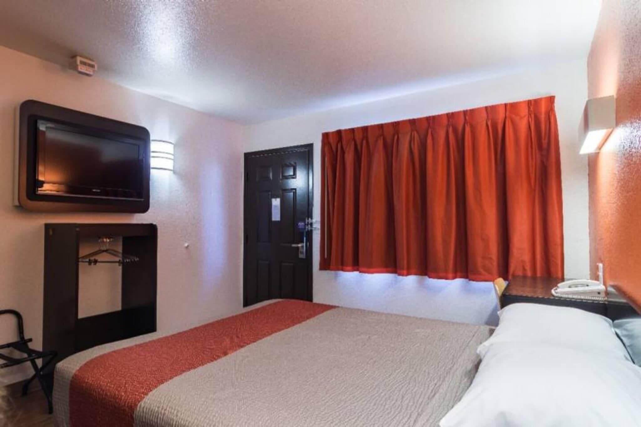 Una habitación de hotel con una cama, un televisor montado en la pared, una puerta y una ventana con cortinas.