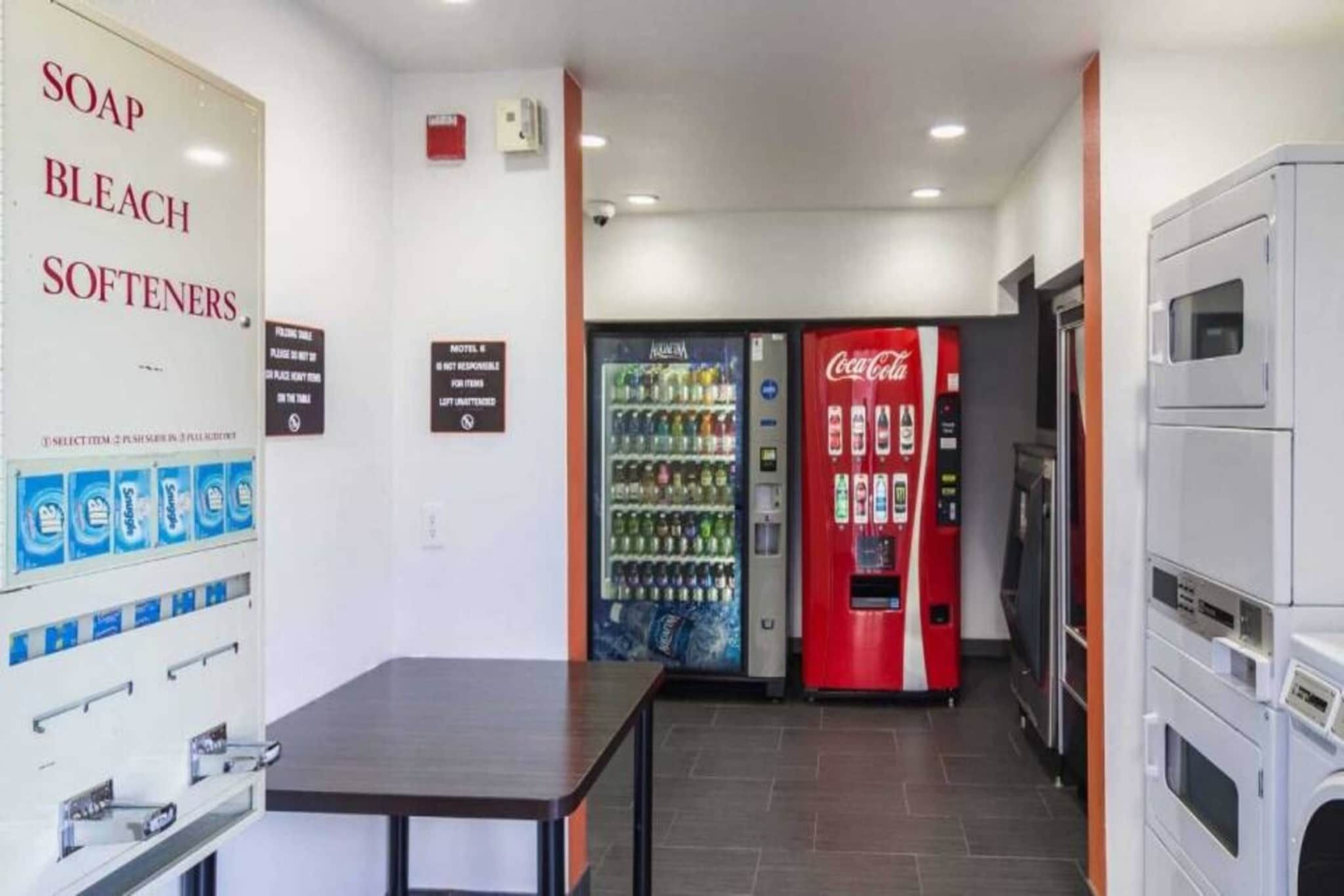 Ein Hotelzimmer mit einem Tisch, einem wandmontierten Spender für Seife, Bleiche und Weichmacher, einer Coca-Cola-Getränkeautomaten und einer Mikrowelle.