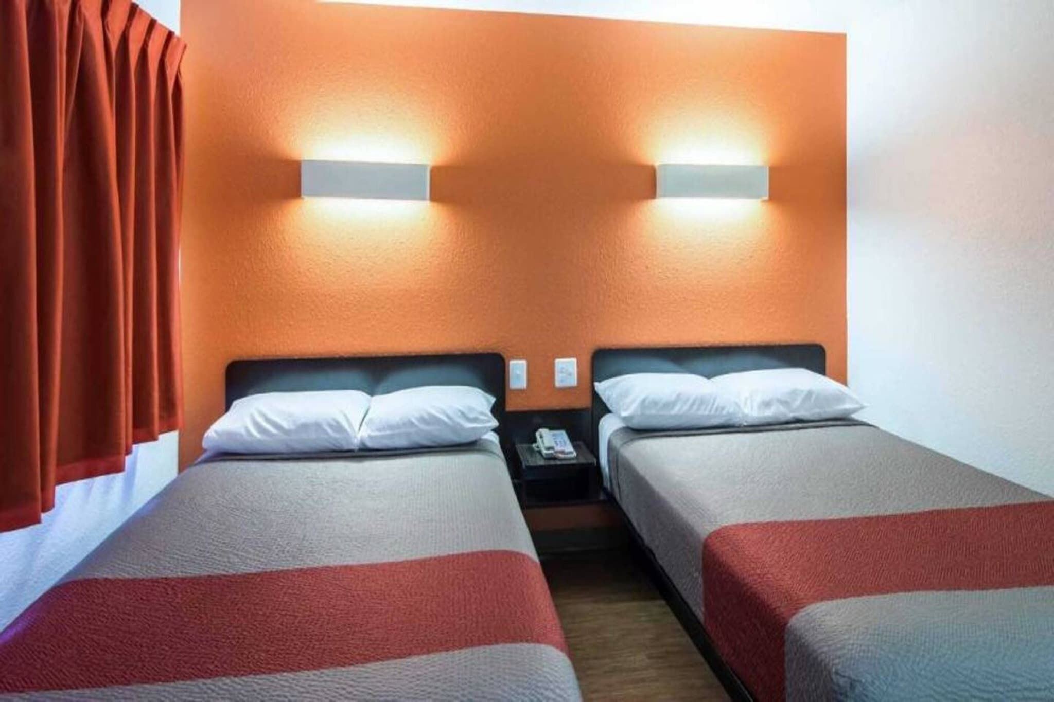 A hotel room with two single beds, a nightstand with a phone, and a wall-mounted light fixture.
