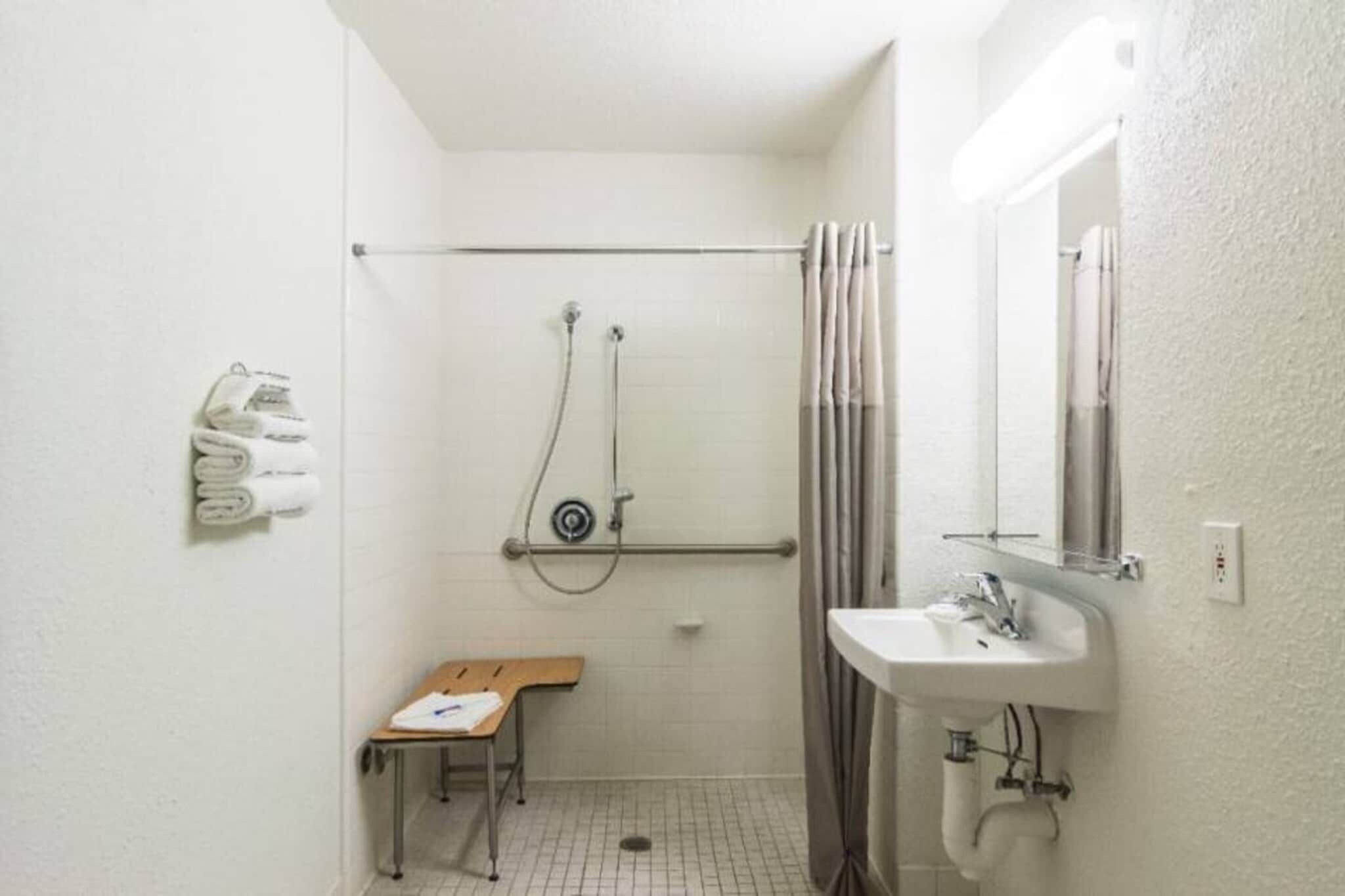 A small, white bathroom with a shower, a sink, and a bench.