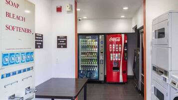 Habitación de hotel con una mesa, dispensador mural para jabón, lejía y suavizantes, máquina expendedora de Coca-Cola y un microondas.