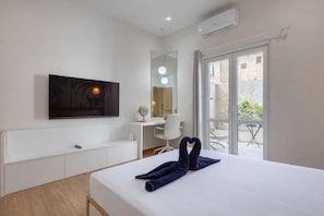 Habitación de hotel con cama, escritorio, silla, televisor y balcón con plantas.
