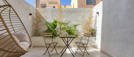 A patio with a wicker chair, a small table, and two chairs. There are potted plants and a wall with a textured surface.