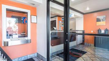 A hotel lobby with a reception desk, a coffee machine, and a seating area with a view through the glass doors.
