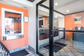A hotel lobby with a reception desk, a coffee machine, and a seating area with a view through the glass doors.