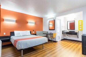 A hotel room with a bed, a desk, a chair, a mirror, and a bathroom with a sink and toilet. - 1 QUEEN BED | Nsmk, MINI FRIDGE (Riverside)