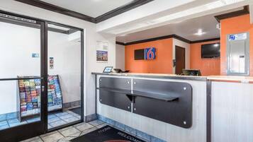 Zona de recepción de un hotel con puerta de cristal, mostrador de recepción, pantalla digital y felpudo de bienvenida.