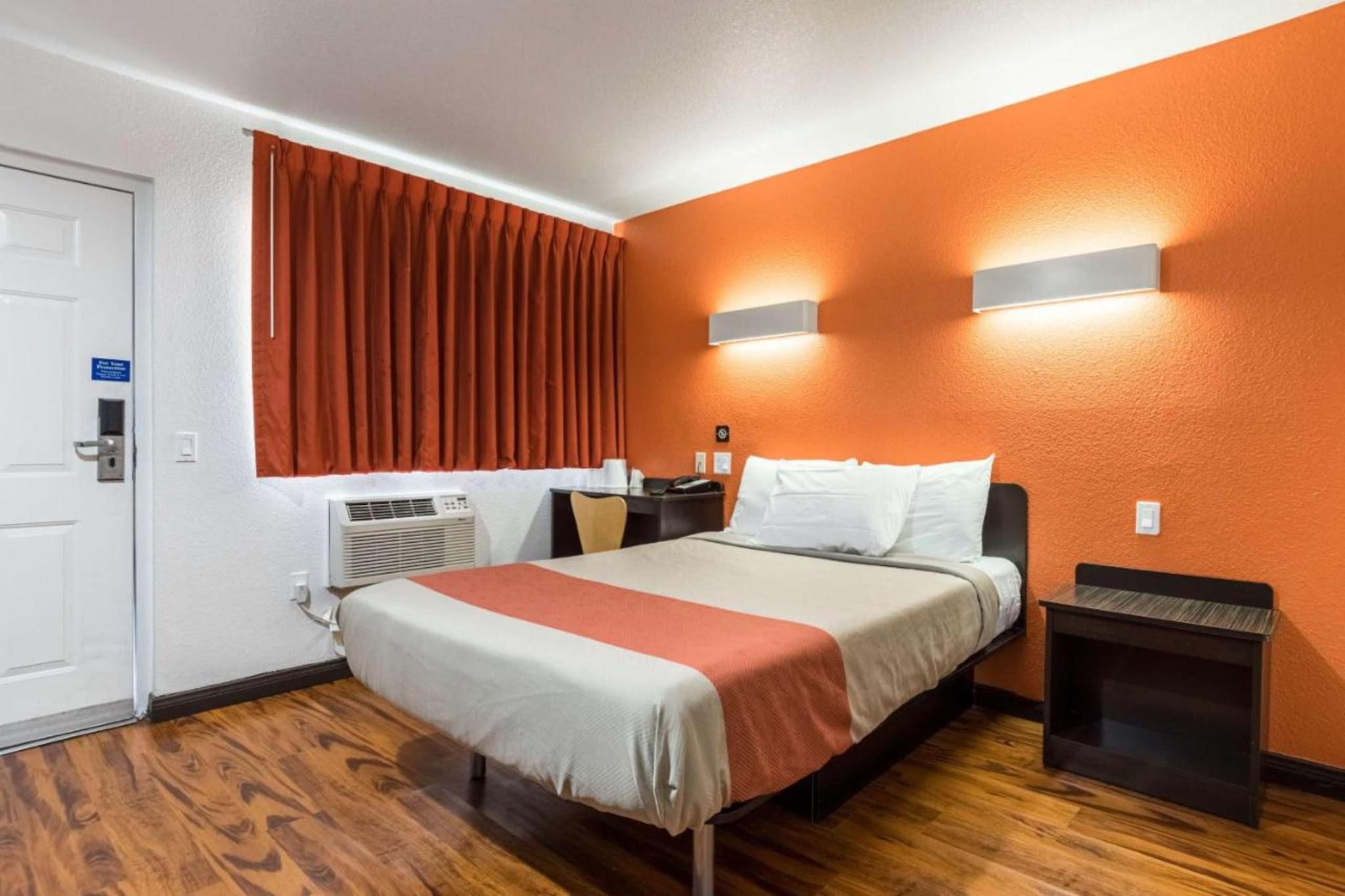 A hotel room with a bed, a desk, a chair, an air conditioning unit, and a door.