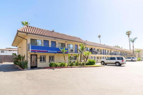 A hotel with a 'WELCOME' sign, multiple floors, and a parking lot with cars. - 1 QUEEN BED | Nsmk, MINI FRIDGE (Riverside)