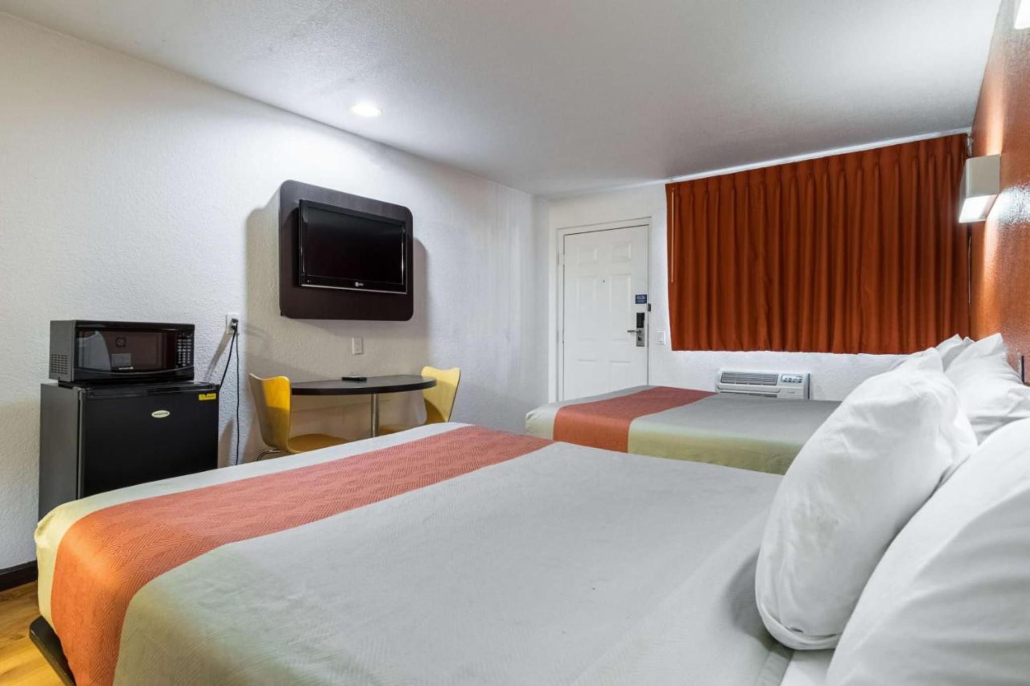 A hotel room with two beds, a desk with a chair, a television mounted on the wall, and a door with a curtain.