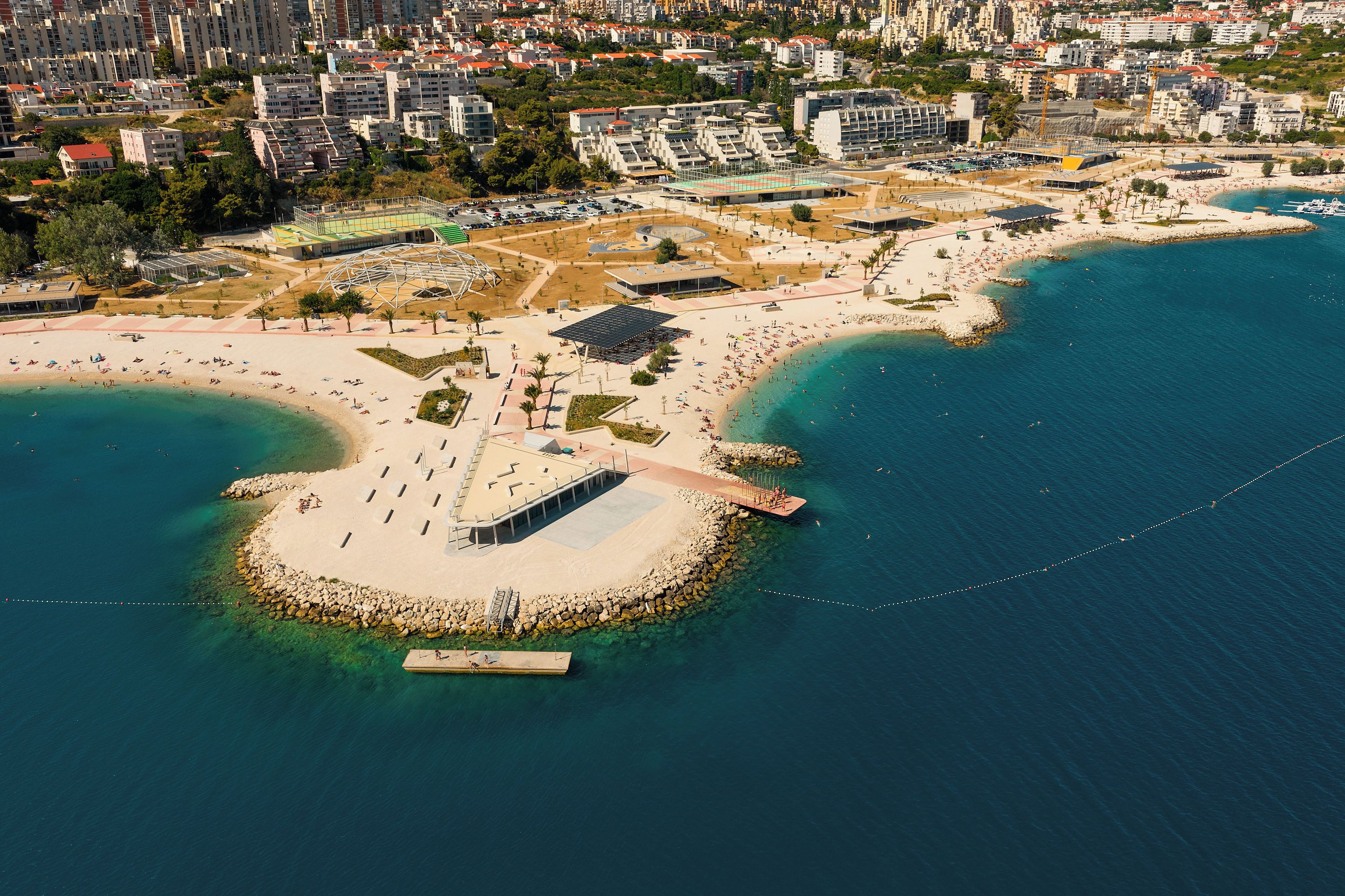 Luftaufnahme einer Küstenlandschaft mit Sandstrand, einer Mole und einer Stadt im Hintergrund.