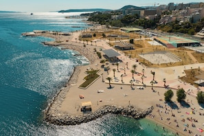Vista aérea de uma área costeira com uma praia, quadra esportiva e estacionamento.