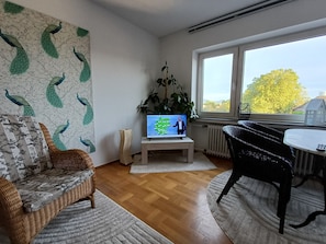 Een woonkamer met een televisie, een plant, een rieten bank, een eettafel en uitzicht op het omliggende landschap.
